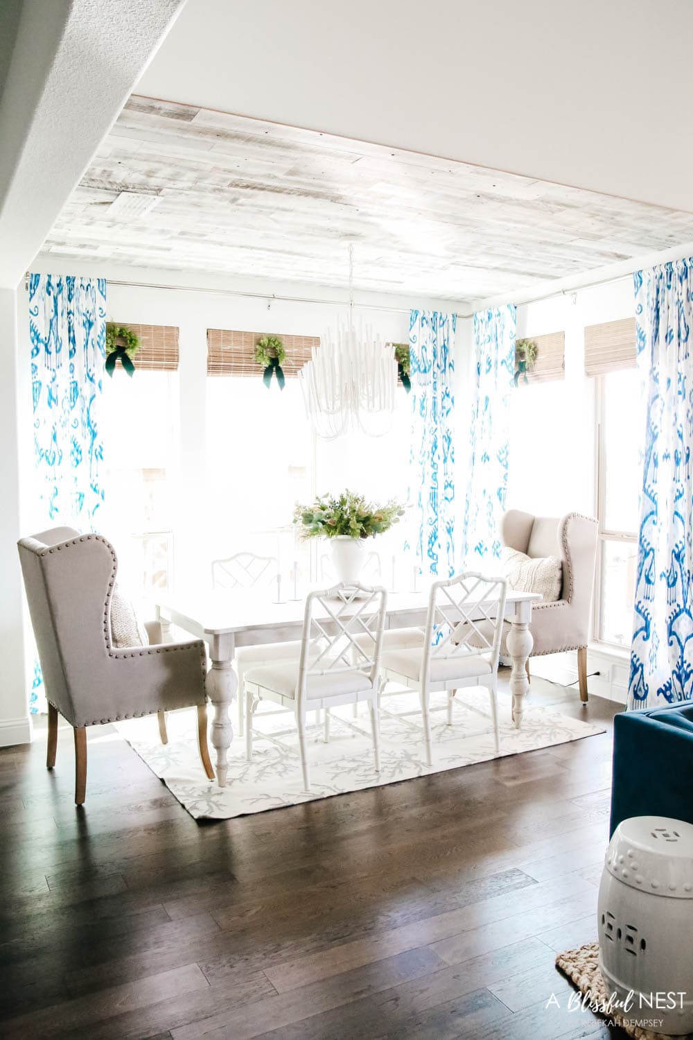 Dining room with white bamboo chairs and blue and white drapes. Simple white candles sticks next to a white vase with juniper floral picks.