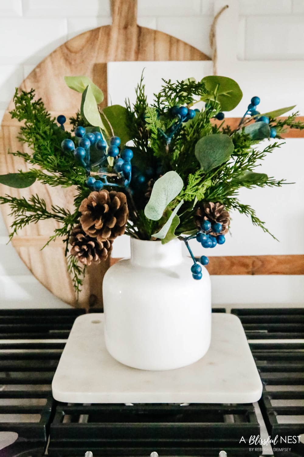 Juniper picks in a white vase sitting on a marble tray on a cooktop in a white kitchen