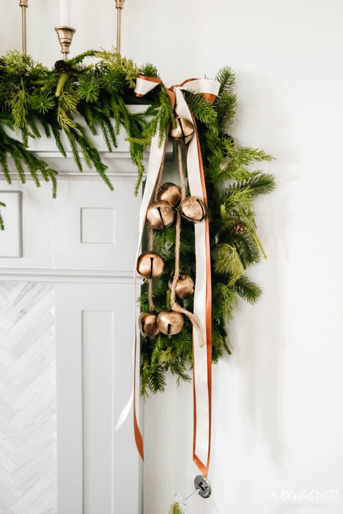 Gold bells and ribbons tied to the side of a garland on a fireplace mantle