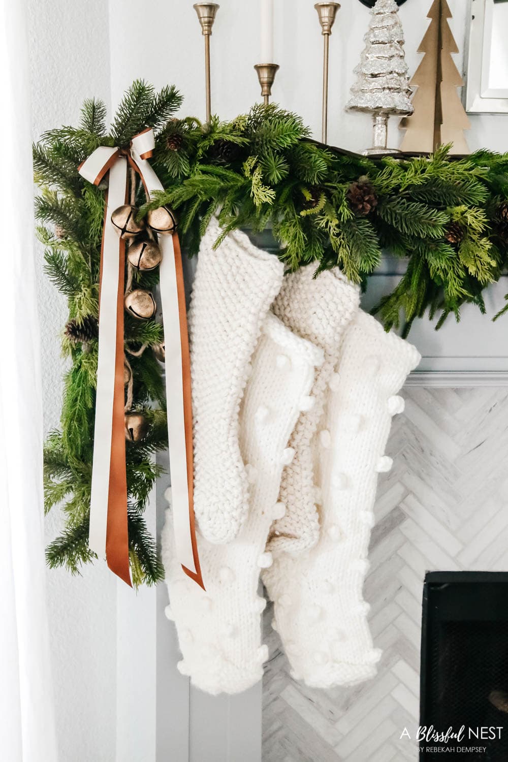 Chunky knit Christmas stockings on the side of a fireplace mantle with garland, gold bells and ribbon