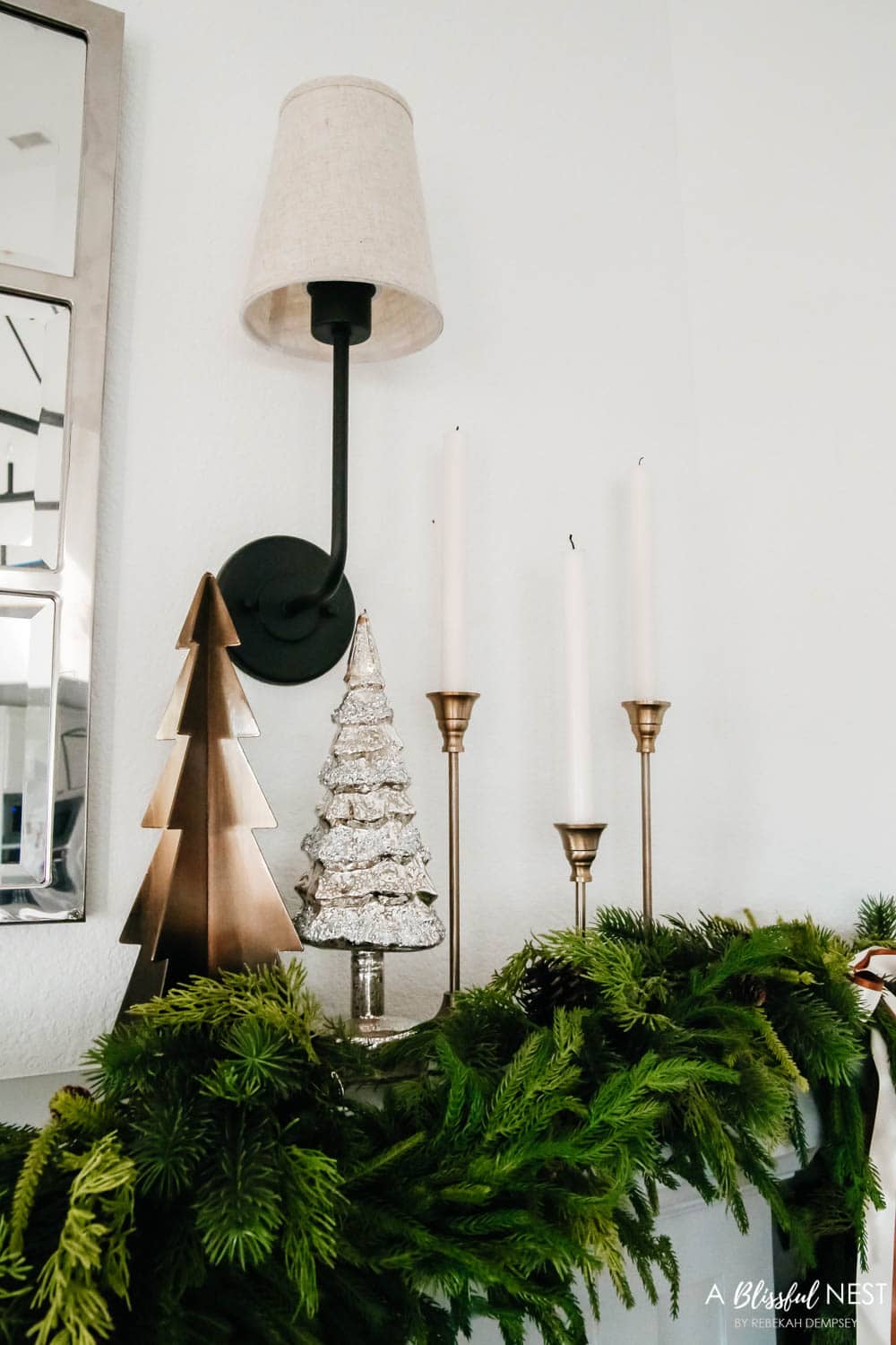 Gold candlesticks, glass and metal Christmas trees with a garland on a fireplace mantle