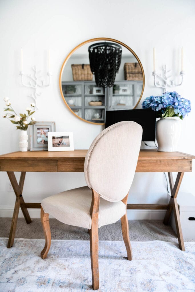 Wood desk with gold round mirror above. Beige tufted side chair used as a desk chair.