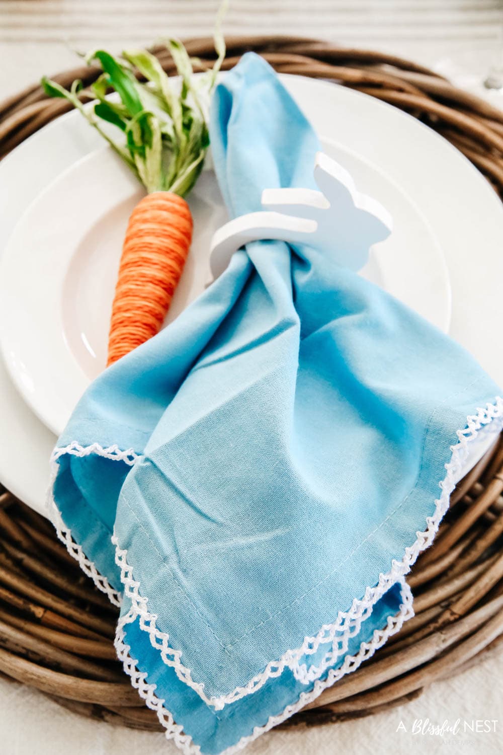 Blue cloth napkin tucked into a white wood bunny napkin holder and a jute carrot resting next to it on a white plate