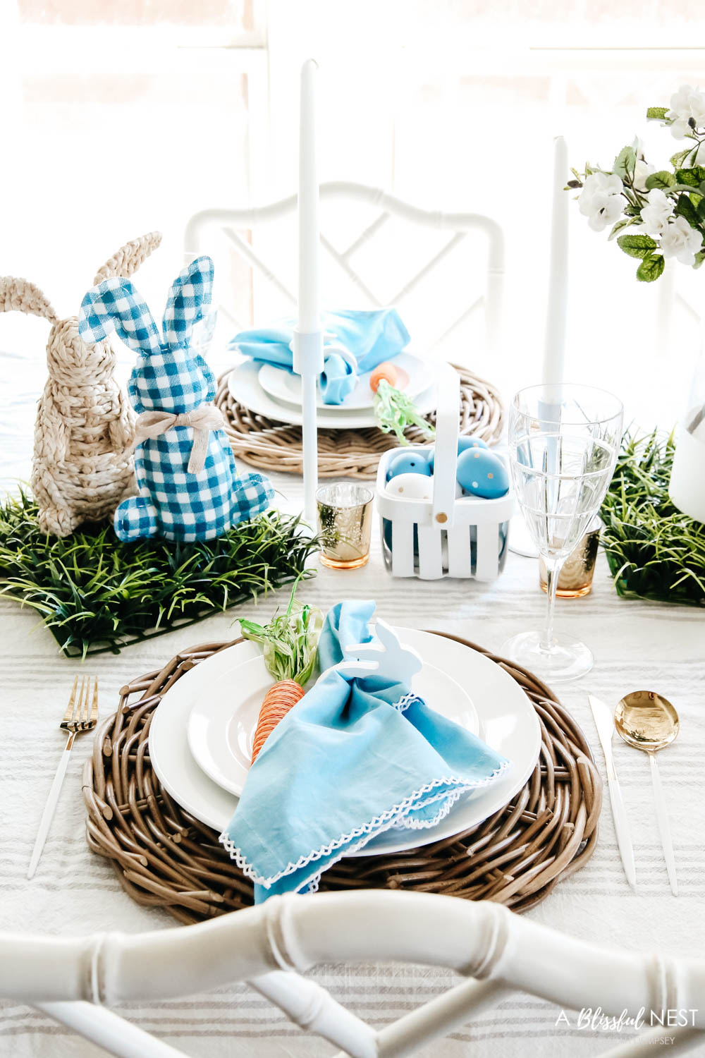 Basket weave placemat with white plates, a blue napkin tucked into a white napkin ring and a jute carrot resting next to it on the plate