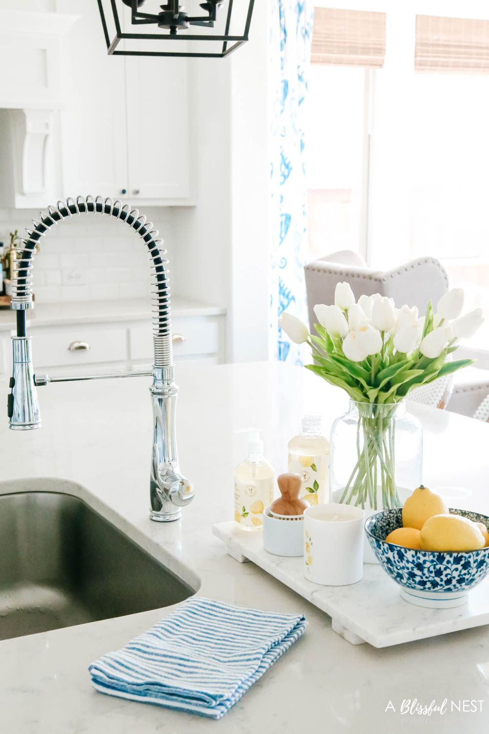 Lemon scented dish soap, scrub brush, bowl of lemons and vase of tulips on a white marble tray.