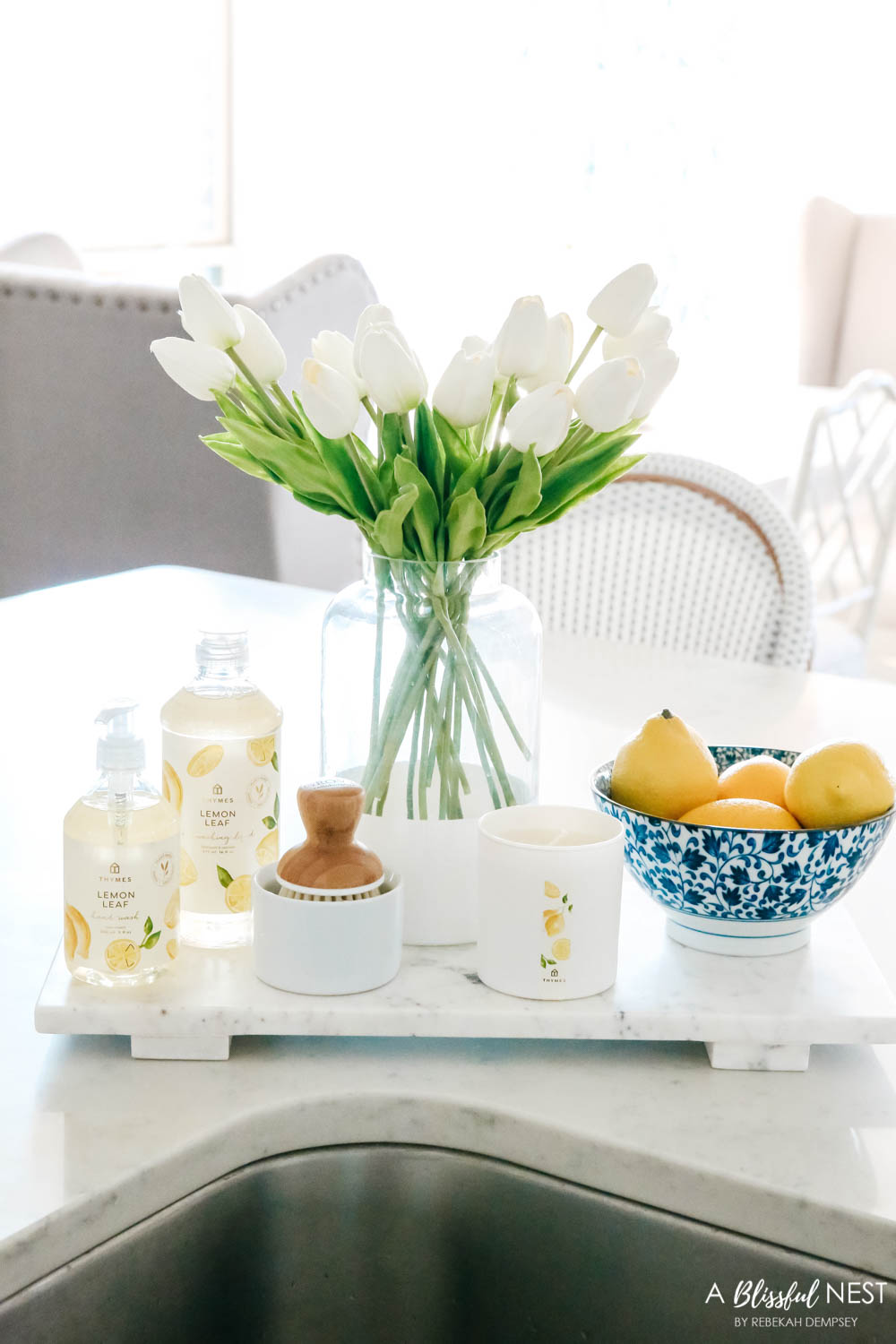 Lemon scented dish soap, scrub brush, bowl of lemons and vase of tulips on a white marble tray.