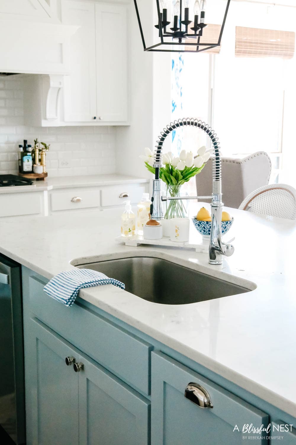 View of kitchen sink with soft blue cabinet color with a vase of white tulips.