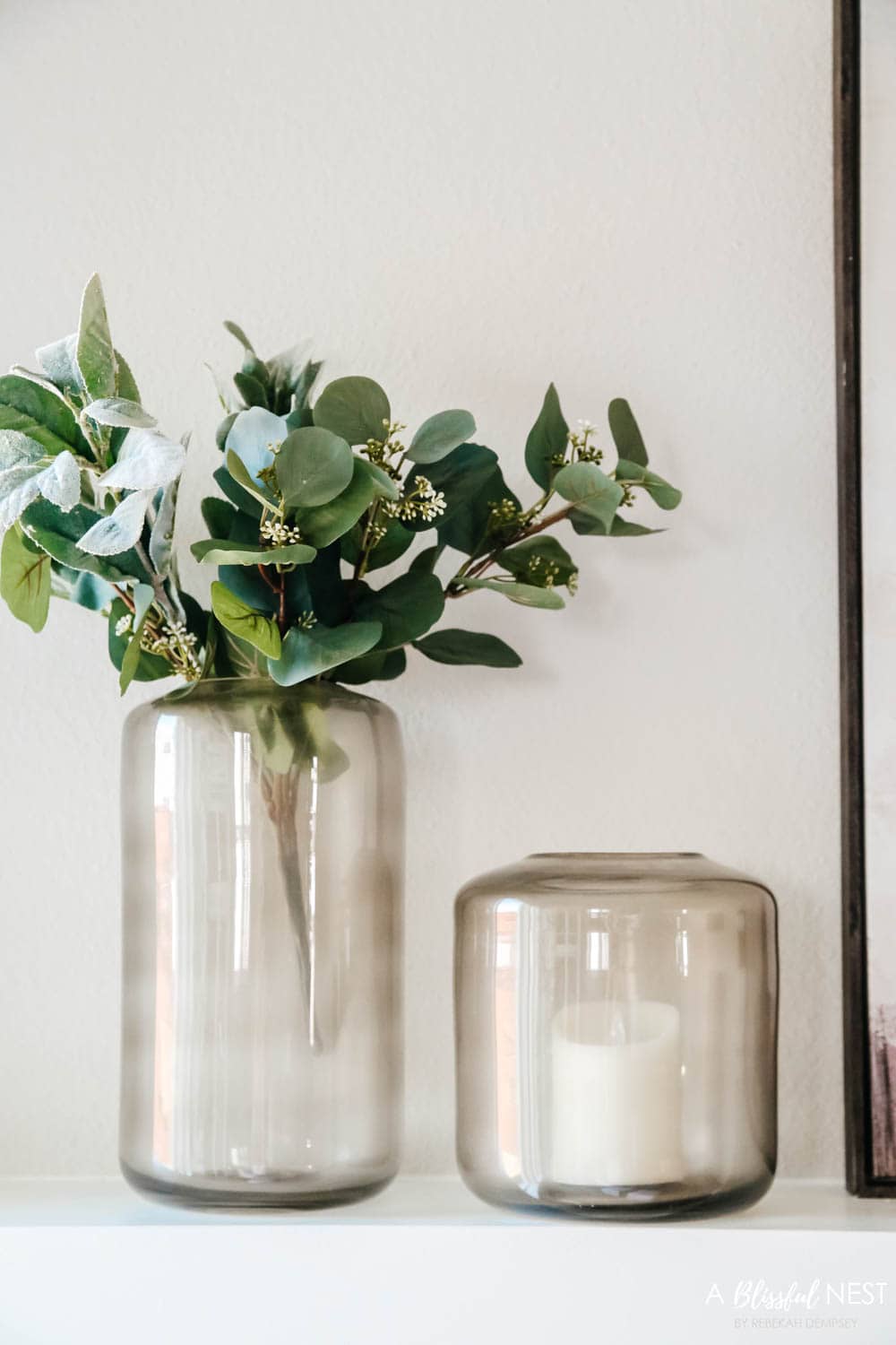 Smoke grey colored hurricane vases with eucalyptus leaves coming out of them.