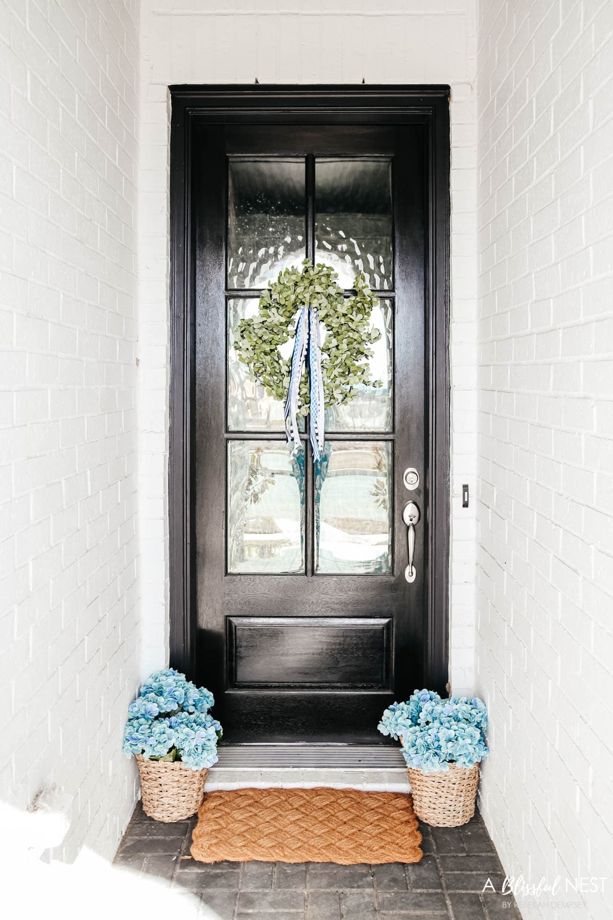 Dark espresso colored front door with eucalyptus wreath with shades of blue ribbon attached to the top, baskets on either side of the door filled with blue hydrangeas