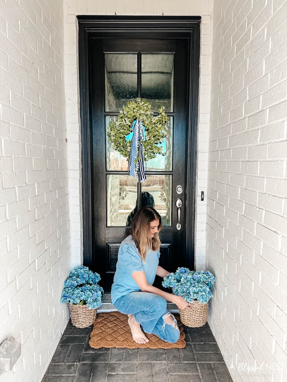 Small porch with a green wreath with ribbons tied to it and baskets of blue hydrangeas 