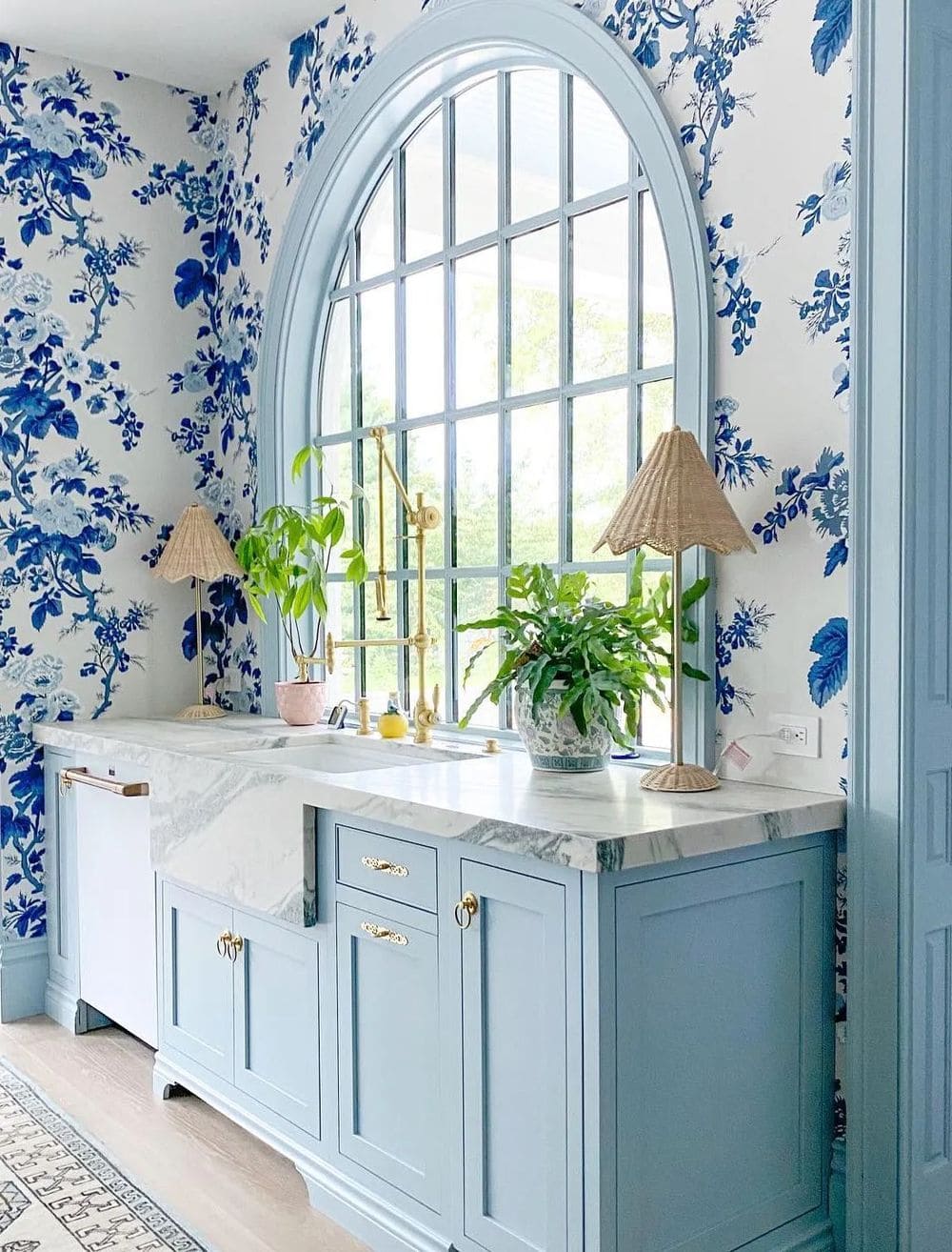 Image of a blue kitchen with blue floral wallpaper. The kitchen features light blue cabinets and white countertops, with the wallpaper adding a decorative touch to the room. The wallpaper has a floral pattern in shades of blue and white, which complements the color scheme of the kitchen. The kitchen is well-lit with natural light, and pots filled with plants sit on the countertop adding a pop of color to the space.