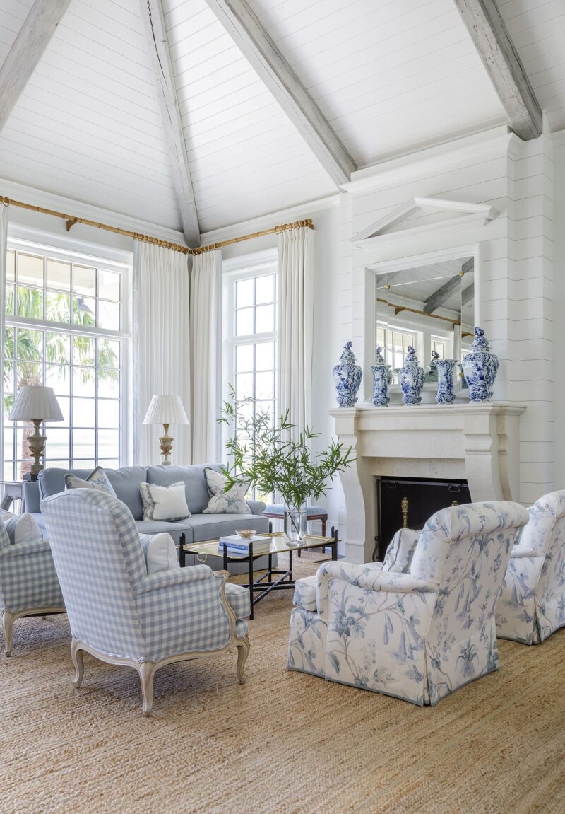 Image of a living room with pale blue patterned chairs and sofa with a sisal rug. The room features two pale blue armchairs and a matching sofa arranged around a coffee table. A sisal rug covers the wooden floor, adding texture to the room. The walls are painted in a white color, with large blue and white ginger jars placed on top of the fireplace mantle. The room is well-lit with natural light from large windows with white curtains.