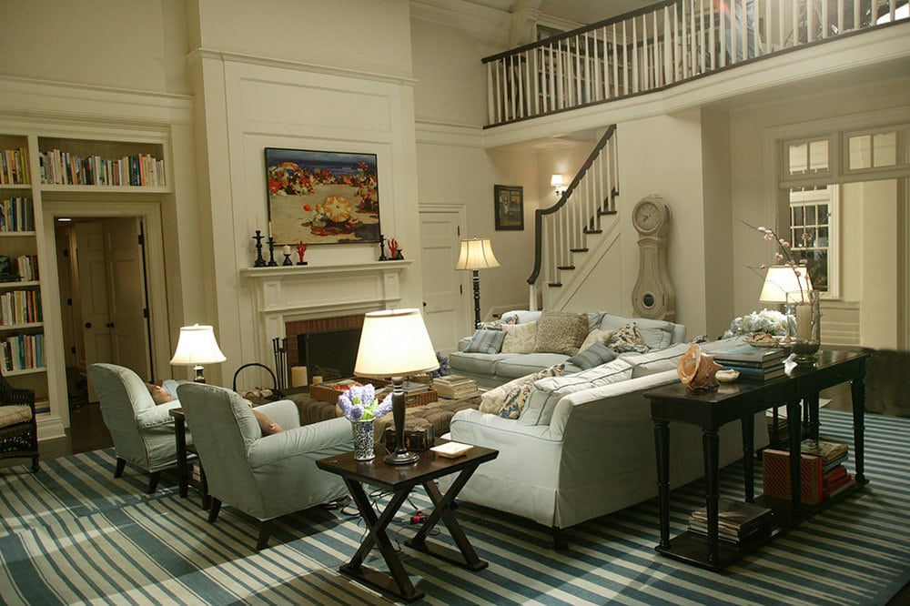 Image of a living room from the movie 'Something's Gotta Give'. The room features a white sofa, several armchairs, and a fireplace. The decor is classic and elegant, with a neutral color scheme of beige and white. A large area rug with a geometric pattern ties the room together, while paintings and decorative items adorn the walls and shelves. The room has large windows that provide natural light.