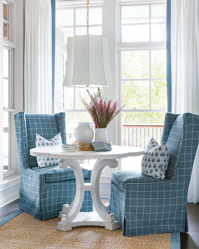 Image of a breakfast room featuring two blue chairs. The chairs are positioned at a round wooden table and have a modern design, with curved backs and metal legs. The chairs complement the blue and white color scheme of the room, which is bright and airy with natural light coming in through the windows. A vase with flowers sits on the table, adding a touch of color to the space.