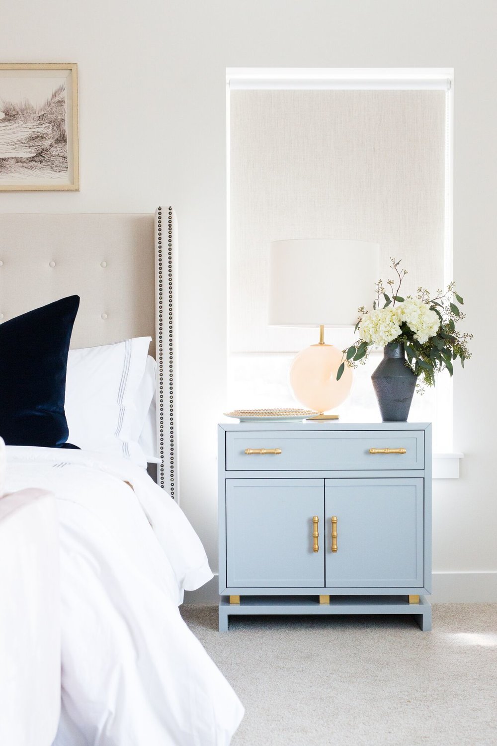 Pale blue nightstand with gold hardware, black vase with hydrangea flowers, table lamp and a bowl.