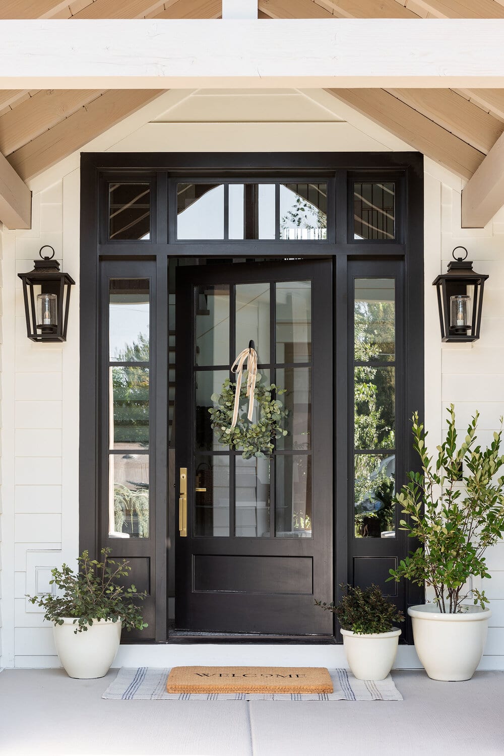 Black front door with black painted side lights. Simple eucalyptus wreath and white potted plants.