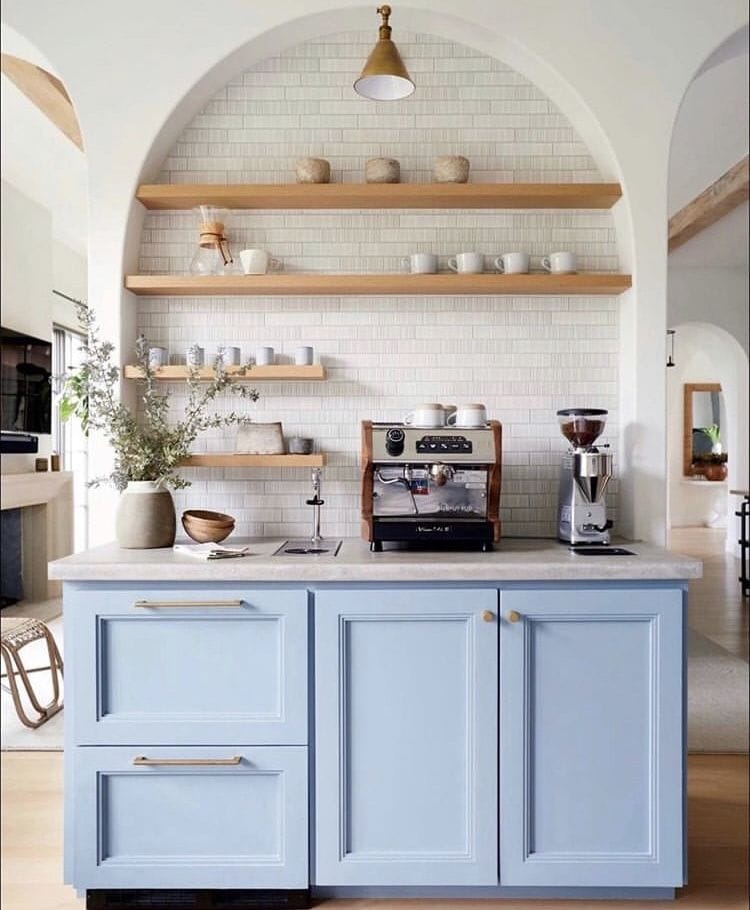 Light blue cabinetry with wood floating shelves above with coffee essentials like a coffee press, coffee mugs, and an espresso machine.