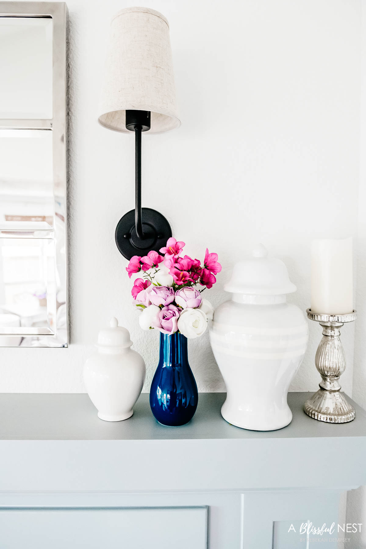 Navy blue vase with purple and pink flowers on a mantle with white ginger jar and mercury glass candlestick.