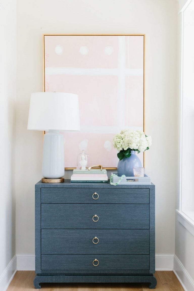 Navy seagrass cloth covered nightstand. Oversized painting behind it with a white table lamp and gold accents.