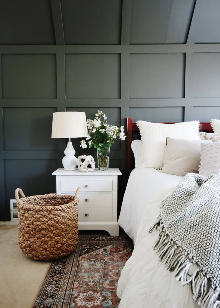 Dark grey painted board and batten on the back of the wall behind the bed with a white nightstand and white table lamp.