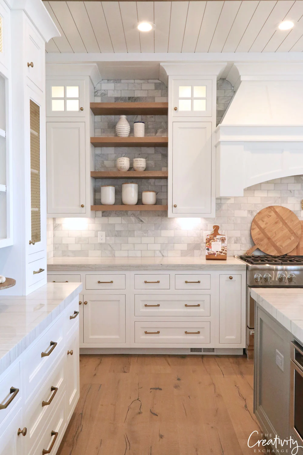 White kitchen cabinetry, gold cabinet hardware, wood floating shelves, wood cutting boards stacked behind range