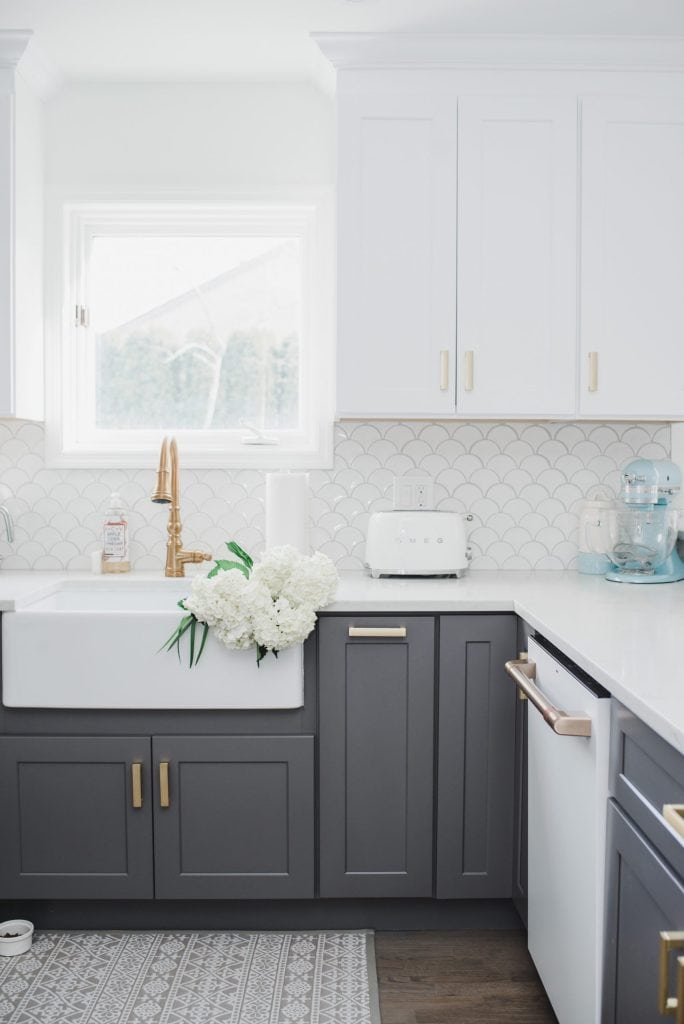Dark grey lower cabinets in a kitchen with white farmhouse sink, gold faucet, white upper cabinets, gold cabinet hardware