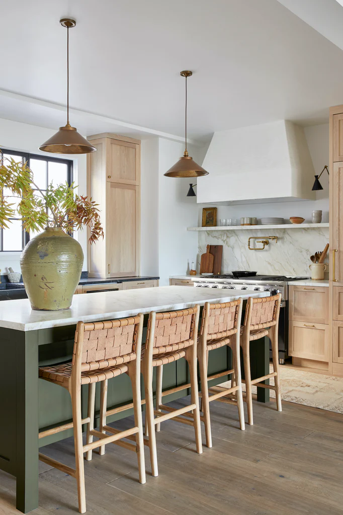 Deep green kitchen island, oak cabinets, marble countertops, woven leather barstools.