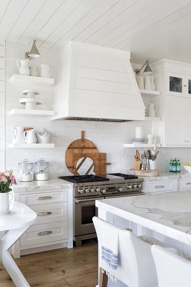 white shiplap hood above range, marble countertops, chrome cabinet hardware, stack of wood cutting boards behind range.
