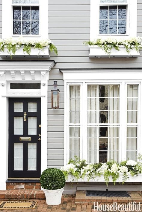Grey siding on a house with white windows and a black door. White window box planters with white flowers.