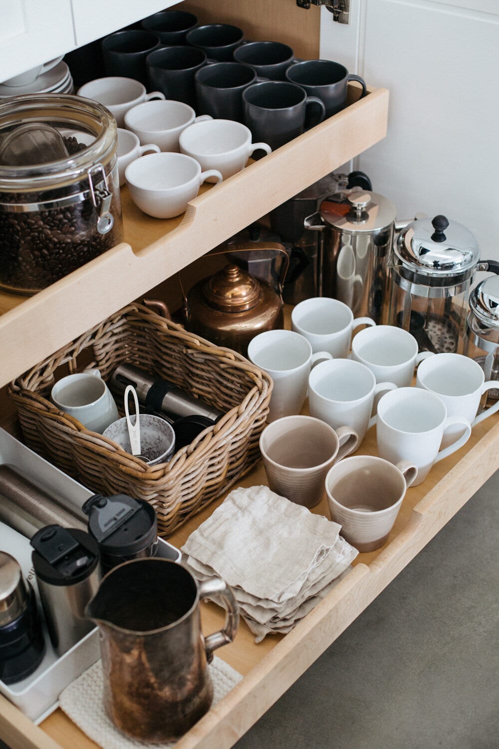 pull out drawer in a kitchen with coffee mugs, jar of coffee beans, french presses, and other coffee tools in a basket