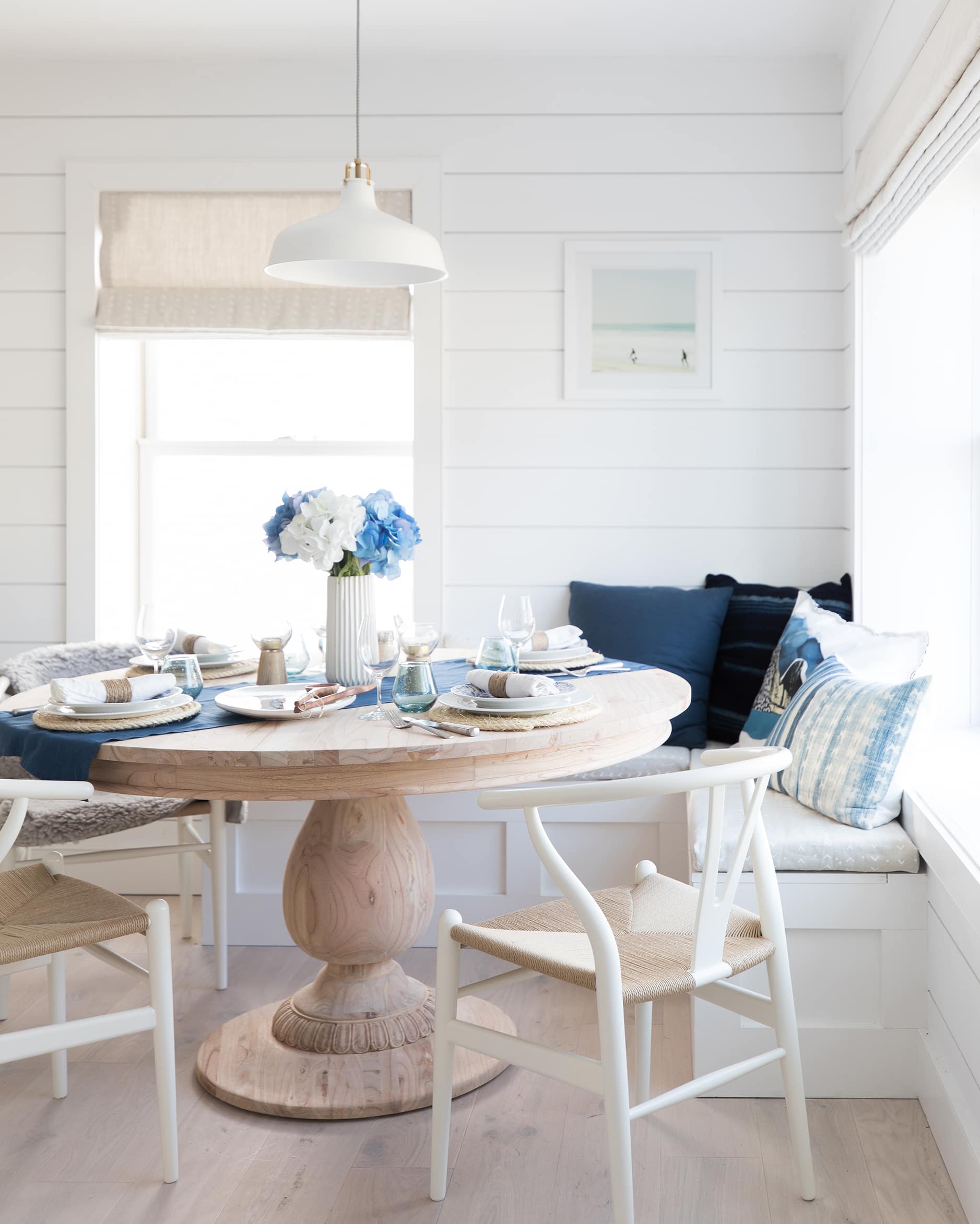 Breakfast nook area with a natural wood round table with white wishbone chairs, navy blue pillows, and white shiplap walls