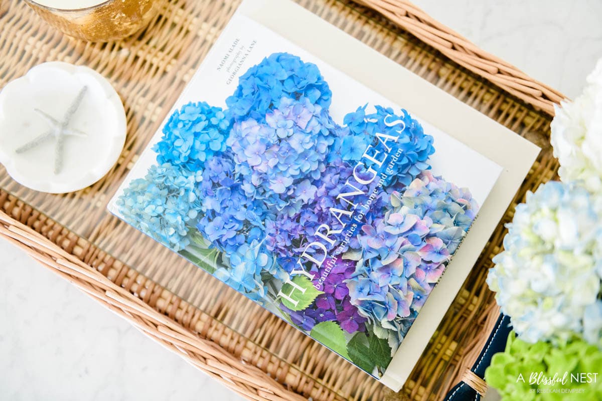 Coffee table book on hydrangeas sitting in a rattan tray on a coffee table.