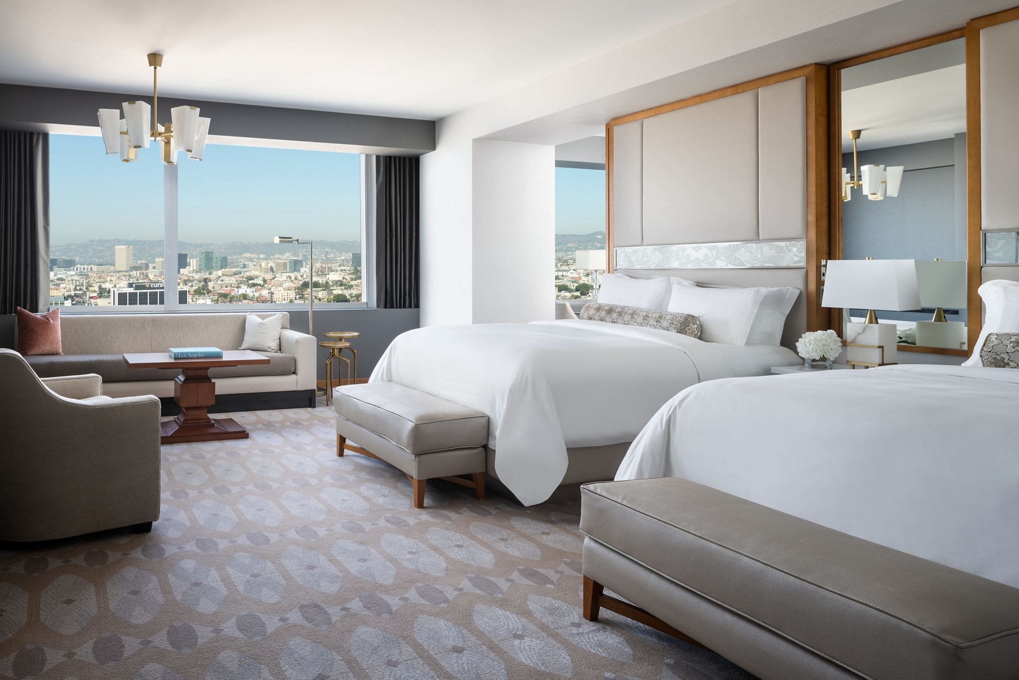 hotel bedroom with all white bedding, grey upholstered bench at end of bed with a city view