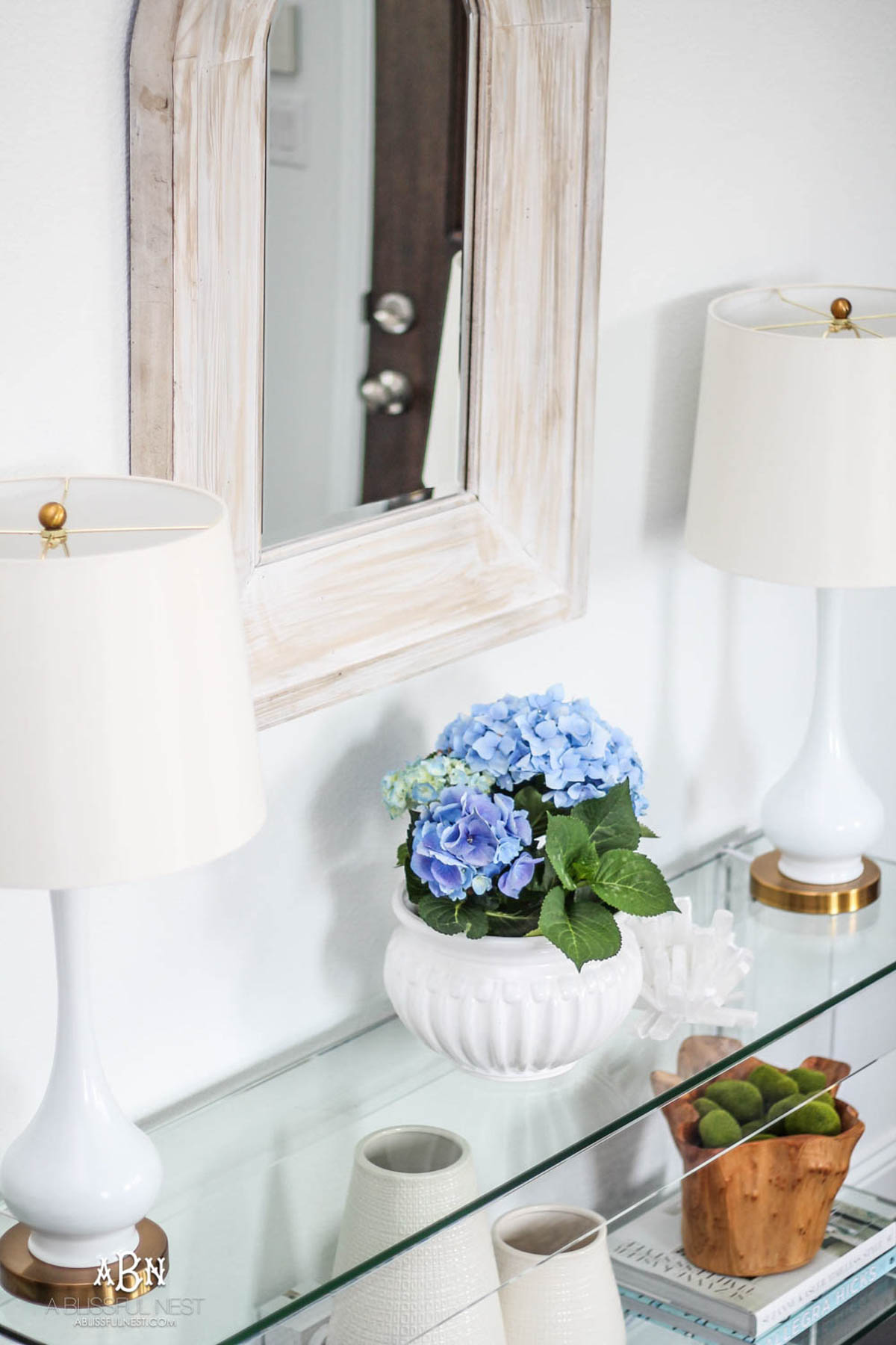 blue hydrangeas in a white ceramic pot next to two white table lamps on a console table.