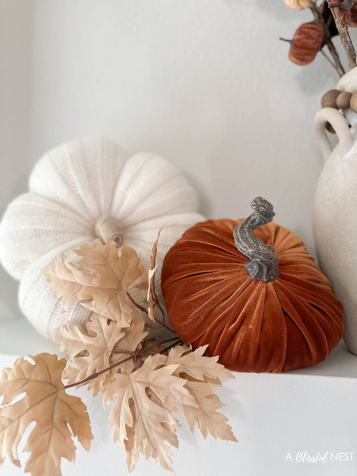 Orange velvet pumpkin clustered next to a cream knit pumpkin with fall leafs tucked into them.