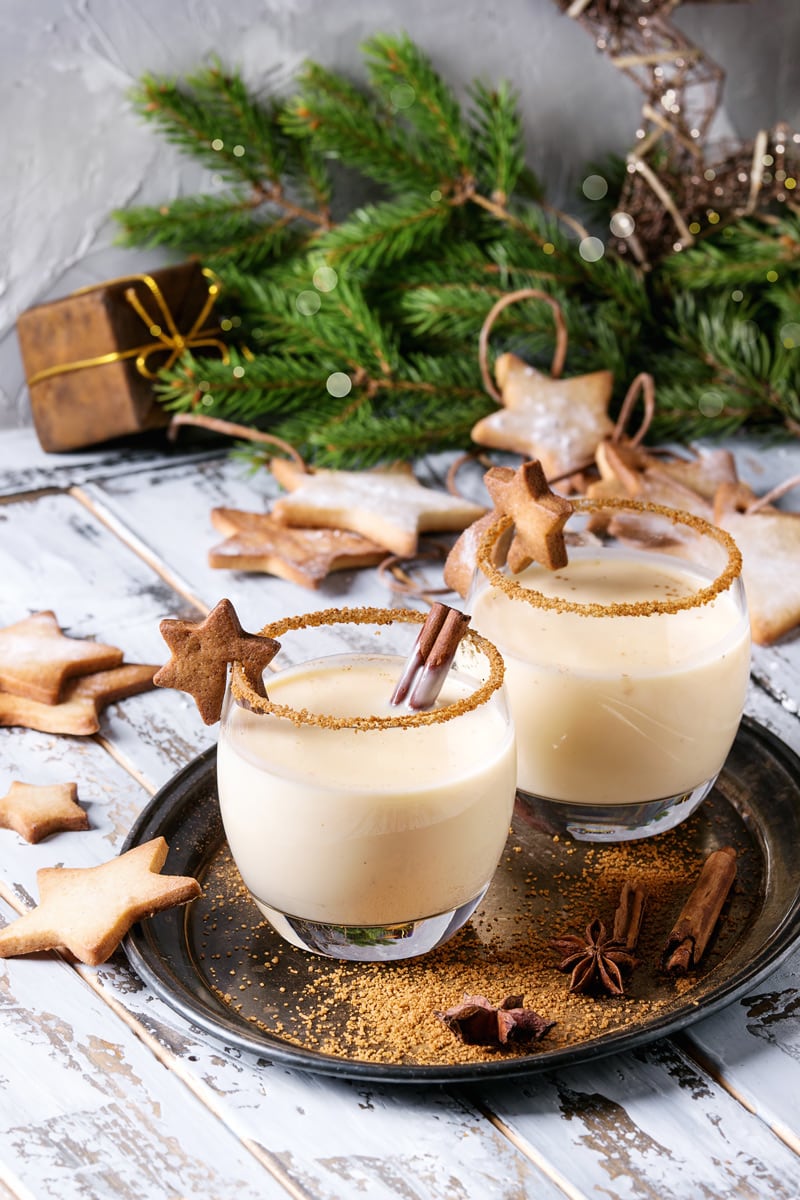 eggnog in a glass with brown sugar on the rim, cinnamon sticks inside, and star gingerbread cookie on rim of drink.