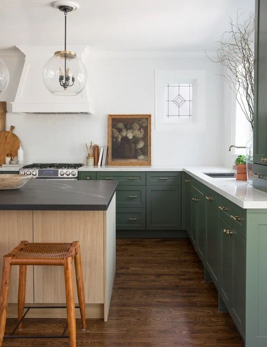 Light wood kitchen island with dark green cabinets for the perimeter. Glass globe lights above the island and a white kitchen hood.